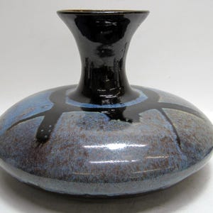 May include: A ceramic vase with a wide, rounded base and a narrow, flared neck. The vase features a glossy black neck and a base with a blue and brown mottled glaze, accented by black linear designs. The vase has a smooth, reflective surface.