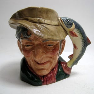 May include: A ceramic mug shaped like an old man's head, wearing a hat. A fish is attached to the side of the mug, forming the handle. The mug is painted in shades of brown, green, and red.