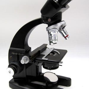 May include: A black microscope with silver and white accents. The microscope has a binocular eyepiece, multiple objective lenses, and a stage for slides. It features adjustment knobs and a mirror for light reflection. A classic scientific instrument.