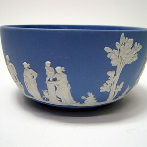 May include: A blue ceramic bowl with white bas-relief figures and foliage. The bowl has a rounded shape and a smooth, matte finish. The white relief depicts classical figures and trees, creating a decorative design.