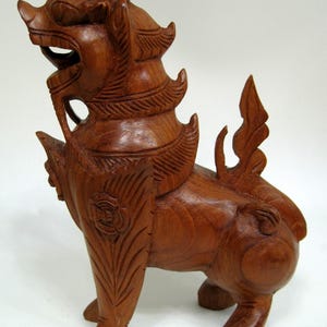 May include: A hand-carved wooden sculpture of a mythical lion-like creature standing in profile. The figure features detailed textures on the mane, wings, and tail, with a smooth, polished brown finish and an open mouth showing sharp teeth.
