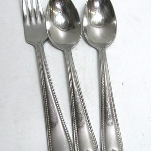 May include: A stainless steel flatware set featuring a fork and two spoons. The utensils have a decorative beaded edge and a stylized floral design at the base of the handles. The polished metal reflects light.