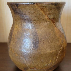 May include: A brown ceramic vase with a wide mouth and a rounded body. The vase has a rustic, hand-thrown look with a mottled glaze.
