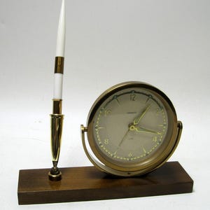 May include: A vintage desk clock and pen set. The clock has a gold-tone frame and a cream-colored face with black numerals. A white pen with gold accents stands to the left. The set is mounted on a dark wooden base.