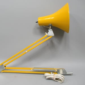 May include: A bright yellow articulated desk lamp with a conical shade and a double-arm metal frame. The lamp features silver-toned metal joints, a coiled white power cord, and a small rectangular label on the base with the text E72400.