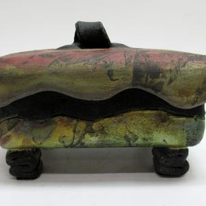 May include: A handcrafted ceramic box with a lid and four feet. The box features a color palette of green, red, and black. The lid has a handle. The box is approximately 6 inches long, 4 inches wide, and 4 inches tall.