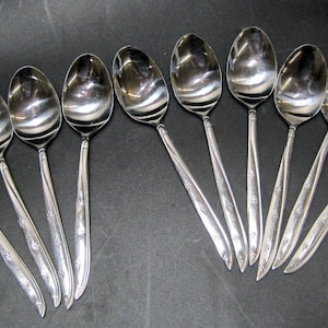 May include: A set of 10 silver spoons with ornate handles. The spoons are arranged in a semi-circle on a black surface.