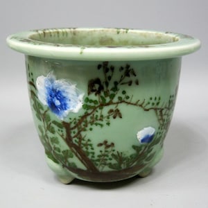 May include: A light green ceramic planter with a wide, flat rim and three small feet. The exterior features a hand-painted design of brown branches with green leaves and prominent blue and white flowers encircling the body of the vessel.
