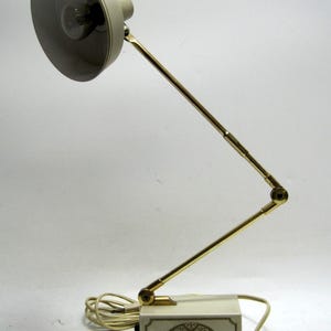 May include: A vintage desk lamp with a cream-colored shade and base, connected by a gold-toned adjustable arm. The lamp has a decorative design on the base and a cord for power. The lamp is approximately 18 inches tall.