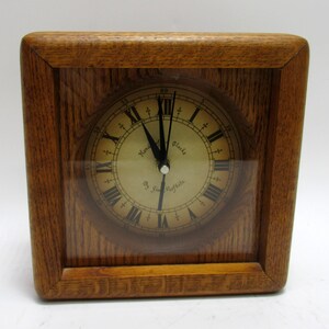 May include: A square wooden clock with a light brown finish. The clock face has Roman numerals and black hands. The text "Handmade Clocks by Jim Moffitt" is visible on the clock face. The clock is encased in a wooden frame.