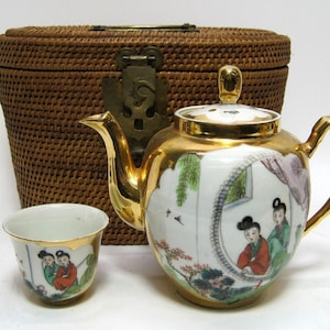 May include: A porcelain tea set with a gold trim and floral designs. The set includes a teapot, a teacup, and a woven basket.