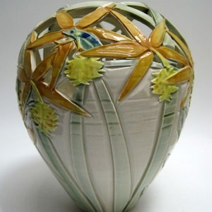 May include: A white ceramic vase with a delicate, openwork design featuring yellow flowers and green leaves. The vase has a rounded, bulbous shape and a smooth, glossy finish.