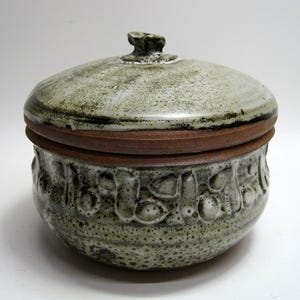 May include: A handcrafted ceramic jar with a lid. The jar is a speckled gray-green color with a dark brown rim and lid base. The jar's body features a textured, embossed pattern. The lid has a small, textured knob for easy handling.