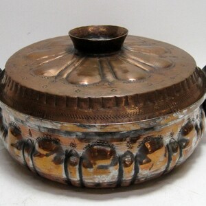 May include: A copper-colored metal cooking pot with a lid. The pot has two handles and a small knob on the lid. The pot's body has a decorative pattern around the base. The lid has a floral design.