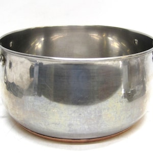 May include: A large, stainless steel cooking pot with two curved handles. The pot has a copper-colored base and a wide opening. The interior is reflective, and the exterior shows signs of use.