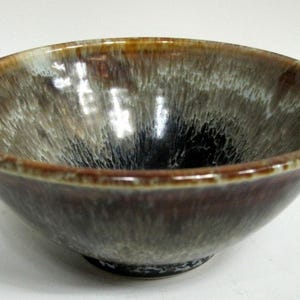 May include: A round ceramic bowl featuring a reactive glaze with a deep black center that transitions into shades of grey, white, and brown toward the rim. The bowl has a smooth, glossy finish and a circular base, showcasing a blend of dark and light tones.