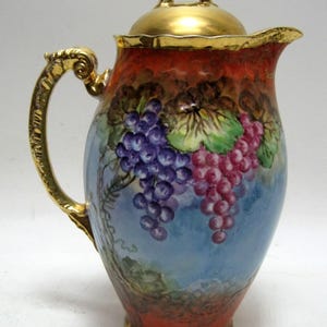 May include: A vintage ceramic pitcher with a gold handle, lid, and trim. The pitcher is painted with a scene of purple and red grapes against a blue and red background. The handle and lid are ornate.