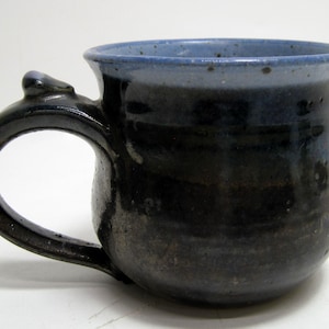 May include: A handmade ceramic mug with a dark blue and black glaze. The mug has a rounded body, a large handle, and a light blue rim. The mug is likely used for drinking coffee or tea.