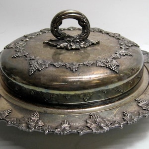 May include: Silver-plated butter dish with a lid and a decorative grape and leaf design. The dish has a round base and a domed lid with a ring handle. The rim of the base and lid are adorned with detailed grape clusters.