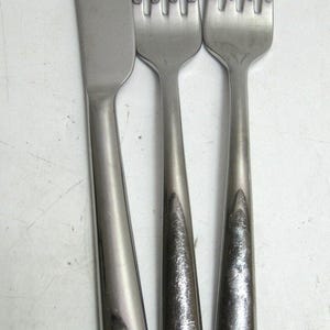 May include: A stainless steel knife and two forks are displayed. The knife has a serrated edge and a long, tapered handle. The forks have four tines each and matching handles. The silverware is set against a white background.