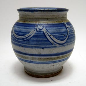 May include: A handmade ceramic vase with a round body and a slightly flared rim. The vase features horizontal blue and white stripes, with a decorative white ribbon-like design around the middle. The base is a darker brown color.