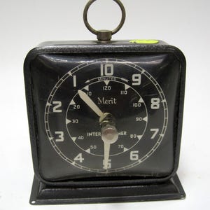 May include: A black Merit timer with a square face and a metal loop at the top. The clock face has white numerals and hands, with the word "Merit" in the center. The timer is set to approximately 10 minutes.