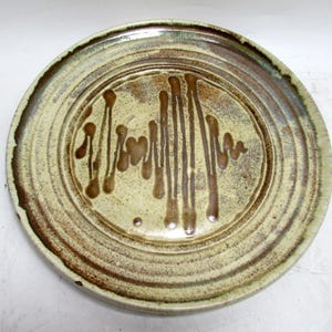 May include: A handmade ceramic plate with a unique design. The plate features concentric rings and a central pattern of brown vertical lines and drips on a beige background. The rim is a darker brown, creating a rustic aesthetic.