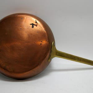 May include: A copper frying pan with a brass handle. The pan is round with a slightly curved edge and a shiny, reflective surface. The handle is long and straight, with a loop at the end for hanging. The pan shows signs of wear.