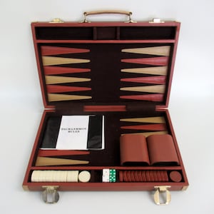 May include: Open backgammon set in a red leather case with a handle. The interior features a dark brown felt playing surface, with tan and red triangles. Includes game pieces, dice, and a booklet titled "Backgammon Rules".