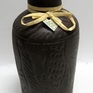 May include: Dark brown ceramic vase with a textured surface and embossed leaf and berry patterns. A raffia bow and tag are tied around the neck. The vase has a wide base and a narrow neck.