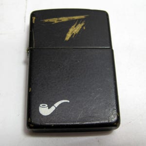 May include: A black lighter with a white pipe graphic on the front. The top of the lighter has a gold-colored design. The lighter shows signs of wear and tear.