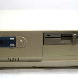 May include: Off-white vintage computer tower with a built-in floppy disk drive and a blue control panel. The front panel has a series of ventilation slots and a small logo. The computer case is rectangular and appears to be from the early 1990s.