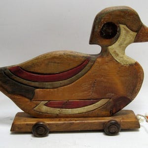 May include: Wooden pull toy in the shape of a duck. The duck is primarily brown with red, black, and cream-colored accents. It has a rectangular base with four small wheels and a pull string with a red bead.