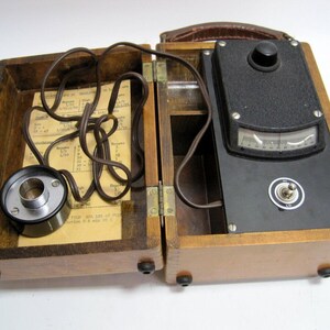 May include: A wooden box with a hinged lid, containing a black electrical device with a dial and a knob. The device is connected to a cord and a metal attachment. The box has a brown leather handle.