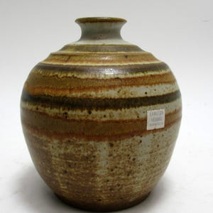May include: A handcrafted ceramic vessel with a rounded body and a narrow neck. The vase features horizontal bands of brown, tan, and cream colors. A small label is affixed to the side. The vessel is suitable for home decor.