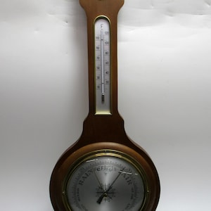 May include: A wooden barometer with a thermometer and a circular gauge. The thermometer displays Fahrenheit readings. The gauge indicates "Rain", "Change", and "Fair". The barometer has a decorative top and a small circular gauge at the bottom.