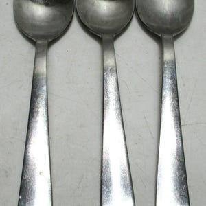 May include: Three stainless steel soup spoons. Each spoon has a rounded bowl and a long, tapered handle. The spoons are arranged side by side, reflecting light. The spoons are likely for kitchen use.