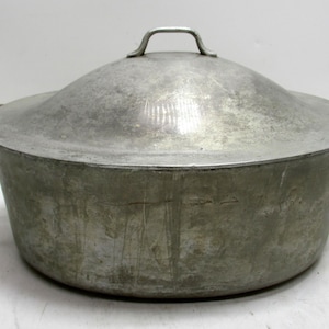May include: A large, vintage aluminum pot with a matching lid. The pot has two side handles and a small handle on the lid. The metal has a dull, aged appearance, suggesting it has been used. This cooking pot is suitable for various kitchen uses.
