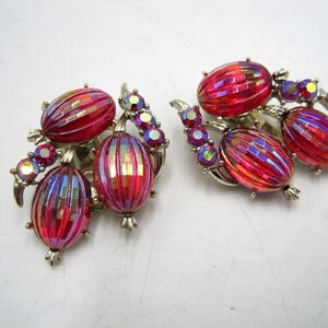 May include: A pair of vintage clip-on earrings. Each earring features three oval, ribbed, red and iridescent glass stones, accented with smaller, iridescent, purple and yellow rhinestones. The earrings have a silver-tone metal setting.