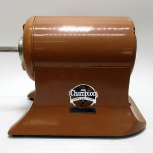 May include: A vintage brown Champion pencil sharpener. The sharpener has a cylindrical body and a base with a curved design. The Champion logo is visible on the front. The sharpener has a silver metal pencil holder.