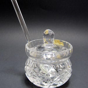 May include: A clear glass honey pot with a lid and a small spoon. The pot has a cut glass design.