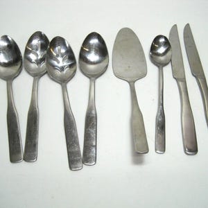 May include: A set of eight silver-toned utensils, including spoons, a serving knife, and a cake server. The utensils have a polished finish and are arranged on a white surface.