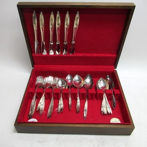 May include: A wooden silverware case with a red velvet interior holds a set of silver utensils. The set includes knives, forks, and spoons, all arranged neatly. The silverware has decorative handles. The case is open, displaying the contents.
