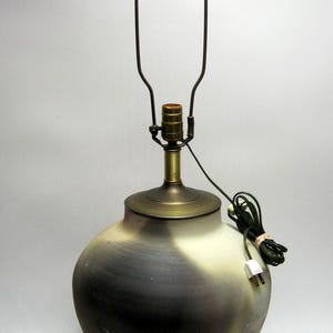 May include: A ceramic table lamp with a bulb holder and a metal shade support. The lamp has a rounded base with a gradient of colors, transitioning from dark gray to cream. The lamp is plugged in.