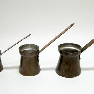 May include: Three copper coffee pots with long handles are arranged on a white surface. The pots are different sizes, with a rounded base and a spout for pouring. The handles are attached to the side of the pots.