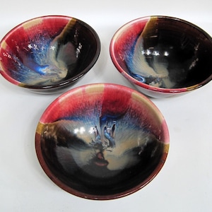 May include: Three ceramic bowls featuring a reactive glaze with a deep black base, transitioning into vibrant streaks of red, blue, and cream. The bowls have a smooth, rounded interior and a glossy finish that creates a swirling, marbled effect.