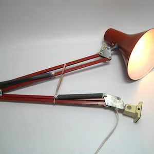 May include: A red and silver articulated desk lamp with a conical shade. The lamp has a pivoting arm design and a cream-colored base. The lamp is extended, and the light is on.