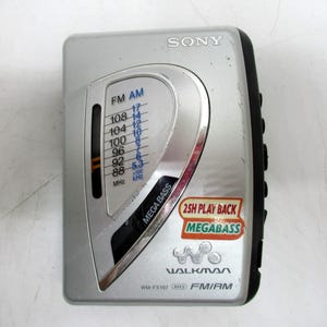 May include: Silver Sony Walkman WM-FX107 FM/AM radio with a black side panel. Features a tuning dial with FM/AM frequency markings and a "Mega Bass" sticker. The Walkman has a 25H Play Back sticker.
