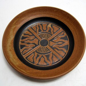 May include: A handmade ceramic plate with a brown rim and a black center. The center features a sunburst design in brown and black. The plate is likely used for serving food or as a decorative piece.