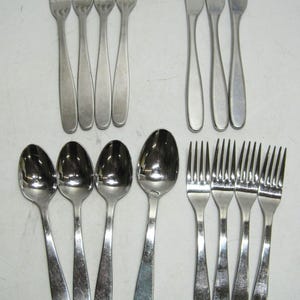 May include: A collection of stainless steel silverware, including forks, knives, and spoons. The forks have four tines, the knives have rounded tips, and the spoons have oval bowls. The silverware is arranged on a light-colored surface.